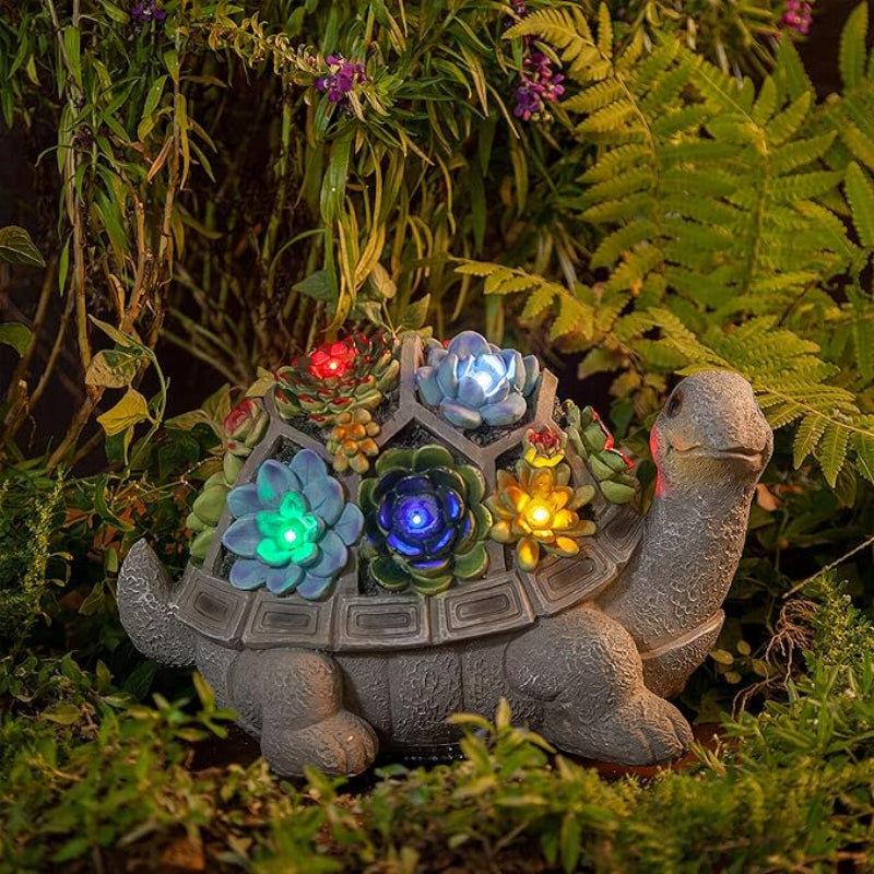 A gray decorative turtle figurine with a segmented shell holding colorful illuminated artificial succulents is placed among lush green foliage and purple flowers outdoors at night. The lights within the succulents emit blue, yellow, and red glows.