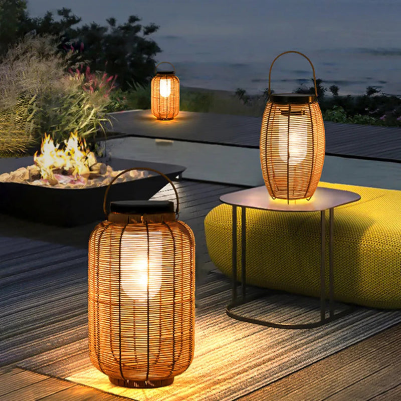 Three warm-toned, cylindrical outdoor lanterns made of woven material are arranged outdoors at dusk. One stands on a wooden surface, another on a small table, and the third on the ground. A fire pit and outdoor seating are visible.