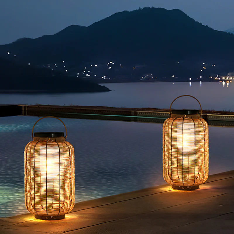 Two warm-toned, cylindrical outdoor lanterns made of woven material emit a soft glow as they sit on a wooden surface at dusk or night. The lanterns have dark tops, possibly solar panels, and metal handles. A body of water with distant lights and a dark mountain are visible in the background.