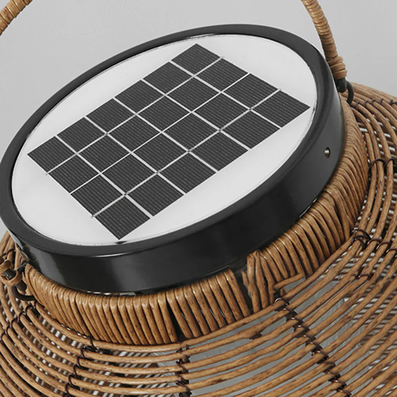 A close-up shows the top of a woven outdoor lantern. A round black cap features a square solar panel with multiple grid lines. A portion of the lantern's woven texture and its handle are also visible.