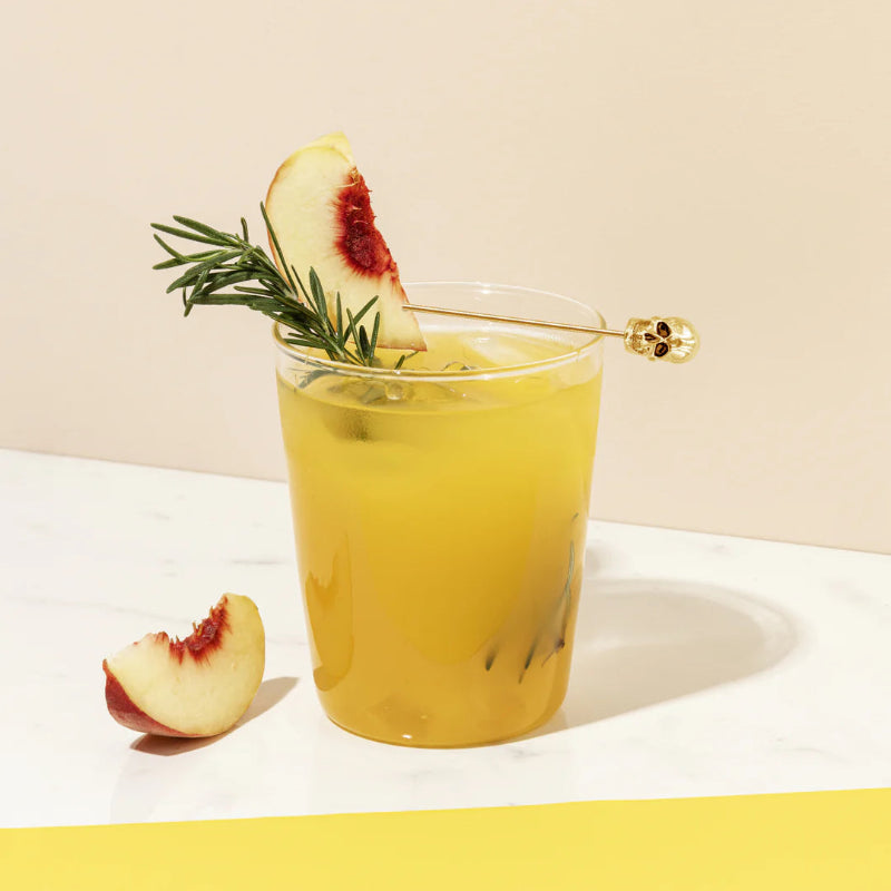 A clear glass filled with a light yellow cocktail, ice, and a sprig of rosemary is garnished with a slice of peach on a gold skull-topped skewer. A separate peach slice rests on the white marble surface next to the glass. The background is split horizontally between a light beige and a vibrant yellow.