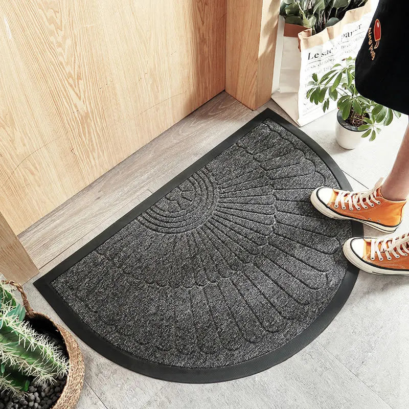 Half-circle grey welcome mat with a feather-like pattern in front of a wooden door, with a person's feet wearing orange sneakers visible.