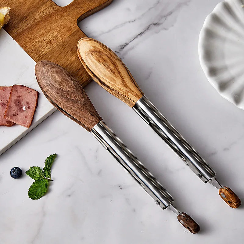 Two stainless steel tongs with wooden handles, one dark and one light, are laid diagonally on a marble surface. Sliced meat, a blueberry, and mint leaves are visible near a wooden cutting board, and a white dish is in the upper right corner.