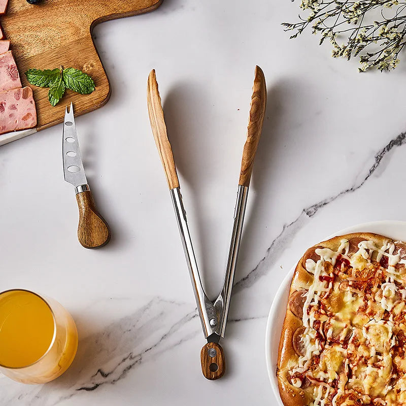 Overhead shot of stainless steel tongs with wooden handles resting on a marble surface.  A cheese knife, sliced meat, a glass of orange juice, a small pizza, and decorative flowers are also visible.