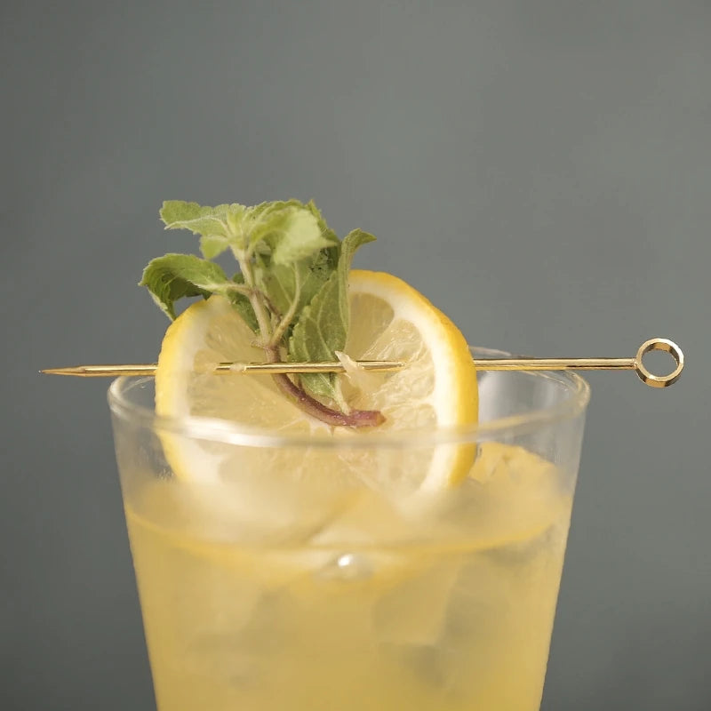 A lemon slice and mint sprig skewered on a gold cocktail pick, resting on a glass of yellow liquid.