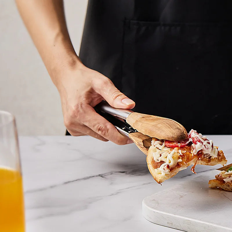 A person wearing a black apron uses stainless steel tongs with wooden handles to pick up a slice of pizza. A glass of orange juice and a marble countertop are also visible.