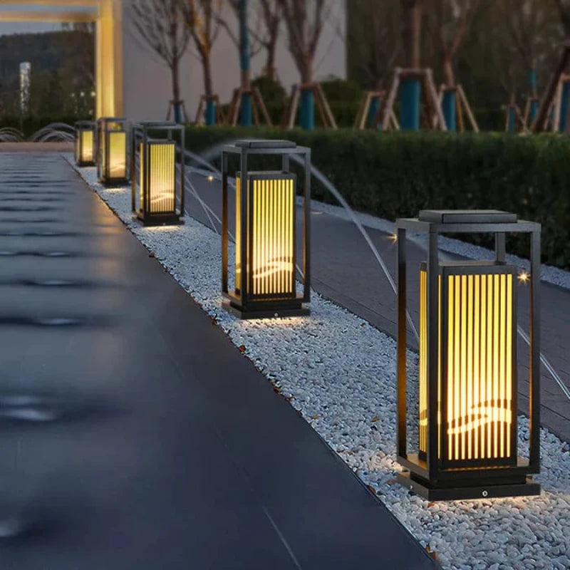 Five modern, dark-framed outdoor lanterns with vertical slats are illuminated along a walkway paved with small white stones at dusk or dawn. The lanterns emit a warm vertical glow, enhancing the pathway. A dark, reflective surface runs alongside the walkway, and blurred trees and buildings are visible in the background.