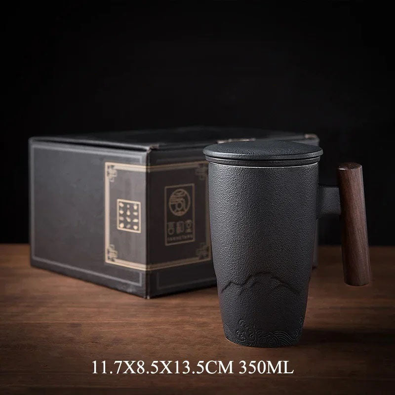 A black ceramic tea mug with an engraved mountain design, a wooden handle, and a matching lid is placed on a wooden surface. A decorative black gift box with gold accents is positioned behind it. The dimensions "11.7 x 8.5 x 13.5 CM" along with the capacity "350ML" are shown at the bottom. The dark background highlights the mug's elegant craftsmanship.