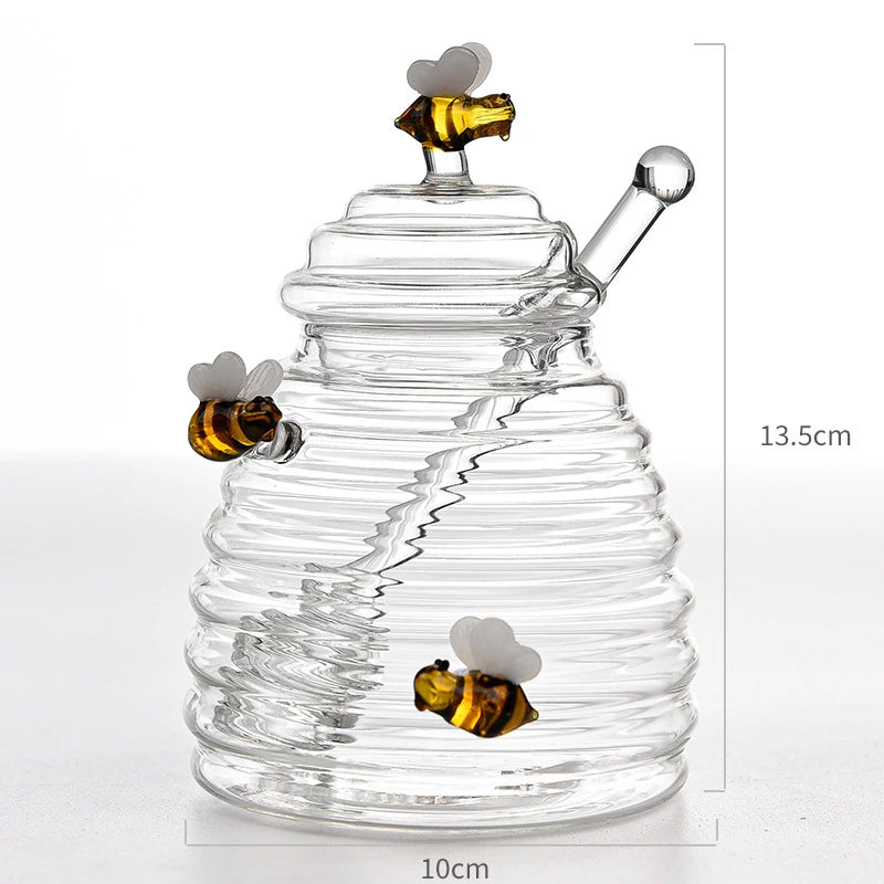 A clear glass honey jar shaped like a beehive with a lid, dipper, and three bee accents, shown with dimensions 10cm width and 13.5cm height.