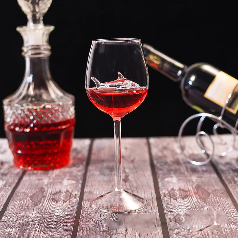 A wine glass filled with a red liquid and a small, clear glass shark figurine inside. A decanter and a wine bottle are in the background.