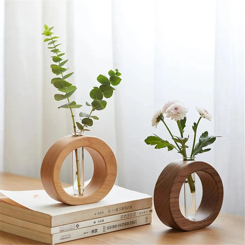 The image shows two circular wooden vases, each holding a transparent glass test tube. One vase is light-colored wood and holds a leafy green stem. The other vase is dark-colored wood and holds white flowers. They are displayed on a light wooden surface.
