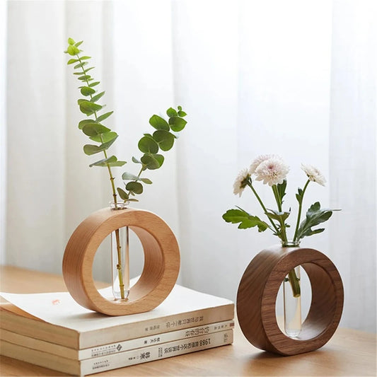 The image shows two circular wooden vases, each holding a transparent glass test tube. One vase is light-colored wood and holds a leafy green stem. The other vase is dark-colored wood and holds white flowers. They are displayed on a light wooden surface.