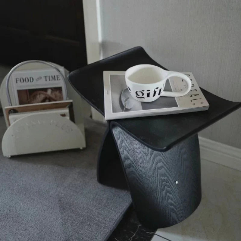 A black Hans Wegner Butterfly Stool sits on a gray rug. A magazine and a white mug with black lettering rest on its top. A white magazine rack with magazines is visible in the blurred background. The floor is partially visible and appears to be marble.