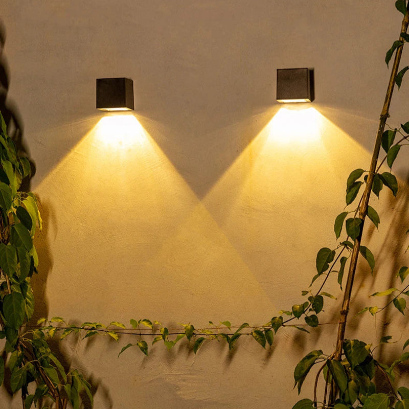 Two black square-shaped wall lights are mounted on a beige wall, casting warm yellow light in a downward triangular pattern. Green climbing plants with vines grow along the wall, adding a natural touch to the illuminated scene.