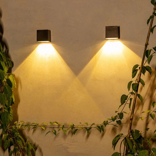 Two black square-shaped wall lights are mounted on a beige wall, casting warm yellow light in a downward triangular pattern. Green climbing plants with vines grow along the wall, adding a natural touch to the illuminated scene.