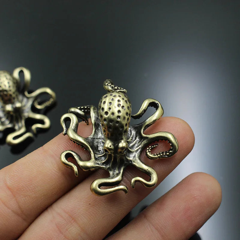 A small antique-style octopus figurine with an intricate design, featuring detailed tentacles and a textured head. The metallic finish gives it a vintage or steampunk aesthetic. It is being held between fingers, showing its compact size, while another identical piece rests on a reflective black surface in the background.