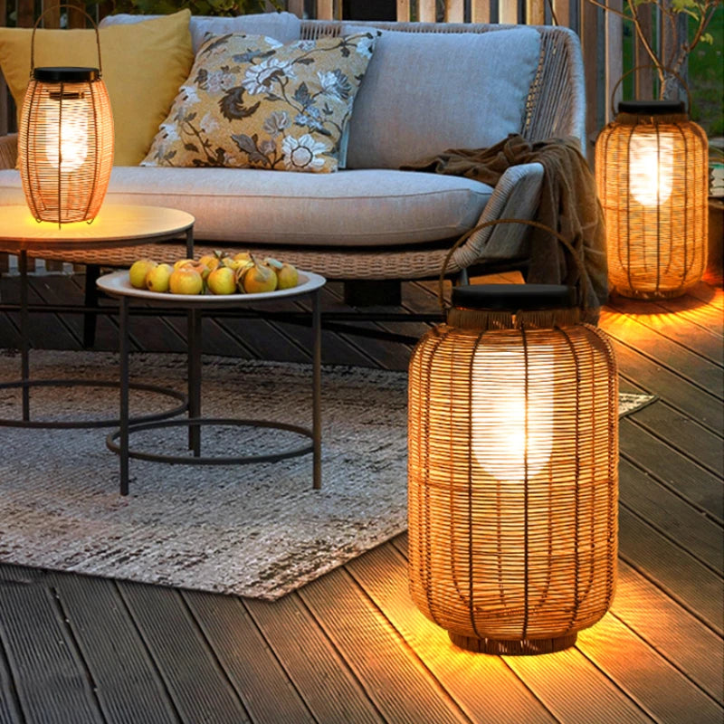 Three warm-toned, cylindrical outdoor lanterns made of woven material emit a soft white glow on a patio at dusk. The lanterns have dark tops, likely solar panels, and metal handles. Outdoor furniture with cushions and a small table with fruit are also visible on the wooden deck and rug.