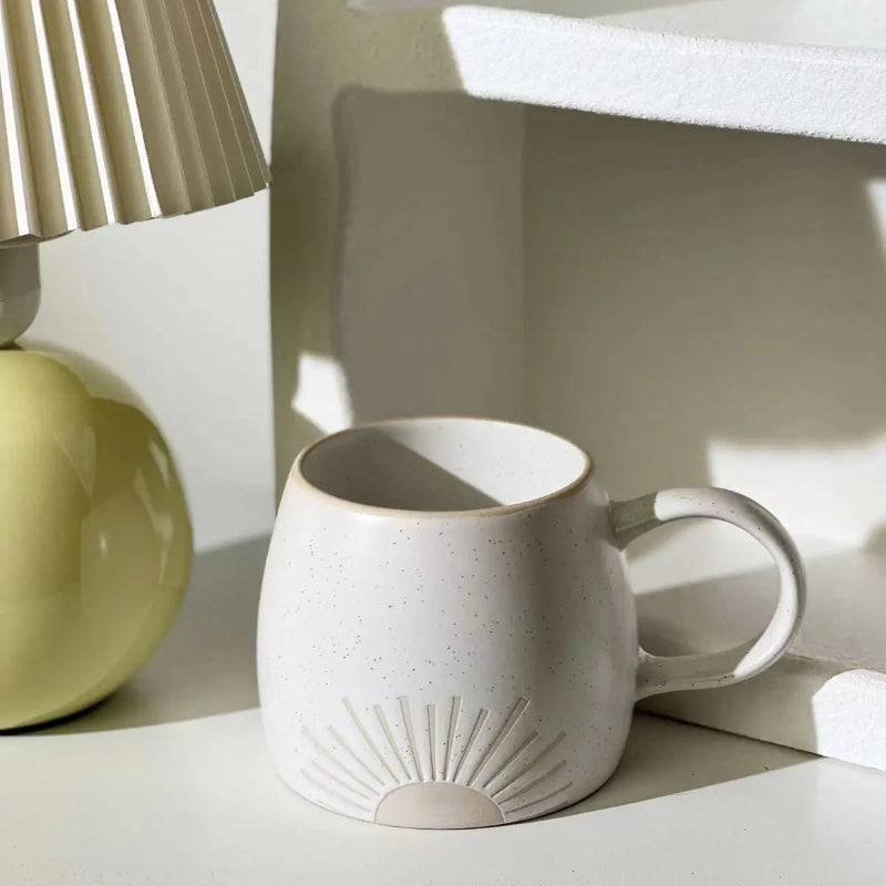 A ceramic mug with a speckled matte glaze and an embossed sunburst design sits on a white surface, illuminated by soft natural light. The mug has a curved handle and an unglazed base. A pale yellow lamp with a pleated shade is placed beside it, adding a warm, minimalist aesthetic to the scene.