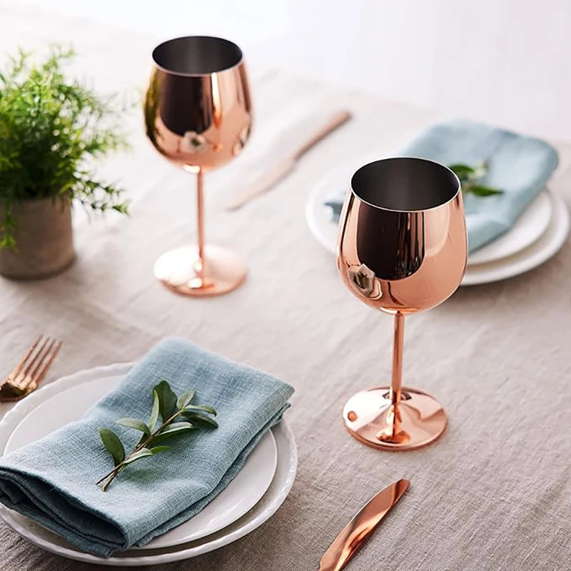 A modern table setting featuring two rose gold stainless steel wine glasses on a neutral linen tablecloth. Each place setting includes stacked white plates, a soft blue napkin adorned with a sprig of greenery, and matching rose gold cutlery. A small potted plant adds a touch of freshness, creating a stylish and inviting dining ambiance.