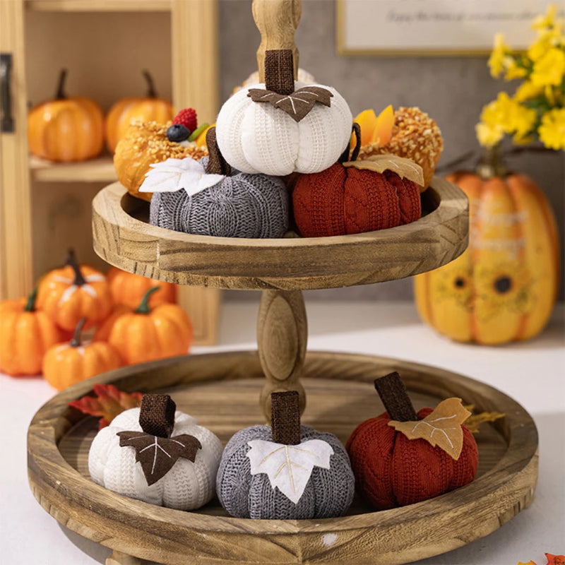 A two-tiered wooden stand holds knitted pumpkins in white, gray, and orange, surrounded by other fall decorations.