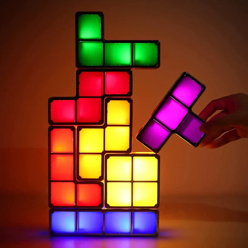 A hand is placing a glowing purple "T" shaped geometric block light onto a partially stacked tower of similar interlocking lights in various colors including blue, yellow, red, and green. The lights emit a soft glow in a dimly lit room.