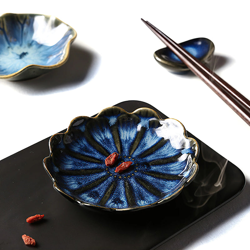 A dark blue, flower-shaped condiment dish with two goji berries, on a black surface next to chopsticks and a chopstick rest.