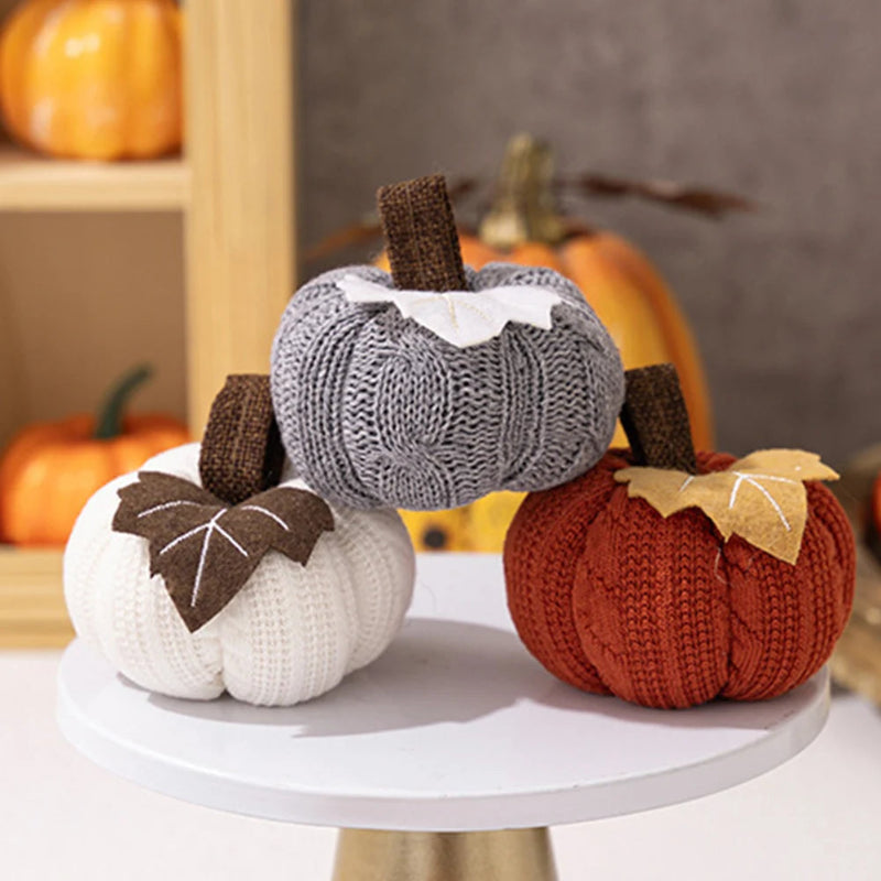 Three knitted pumpkins, in gray, white, and orange, are arranged on a white cake stand.