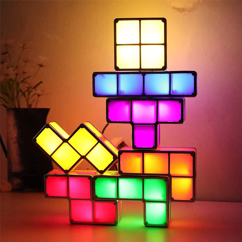 A stack of interconnected, glowing geometric block lights in various shapes and colors, including yellow, blue, purple, red, and green, creating a colorful illuminated structure. The lights are arranged on a surface in a dimly lit room, with a blurred plant visible in the background.