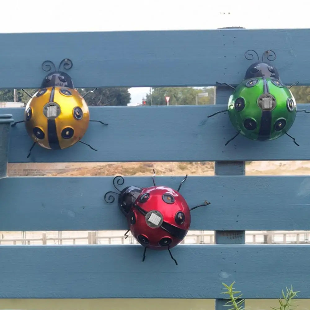 Three decorative metal ladybugs, one yellow with black stripes, one green with black spots, and one red with black spots and stripes, are mounted on a blue wooden fence. Each ladybug has black antennae and legs.