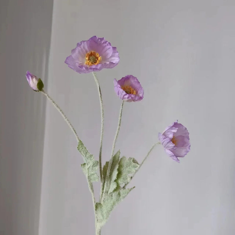 Three lavender poppy flowers with green stems and one poppy bud are arranged in a sparse bouquet against a plain light grey background.