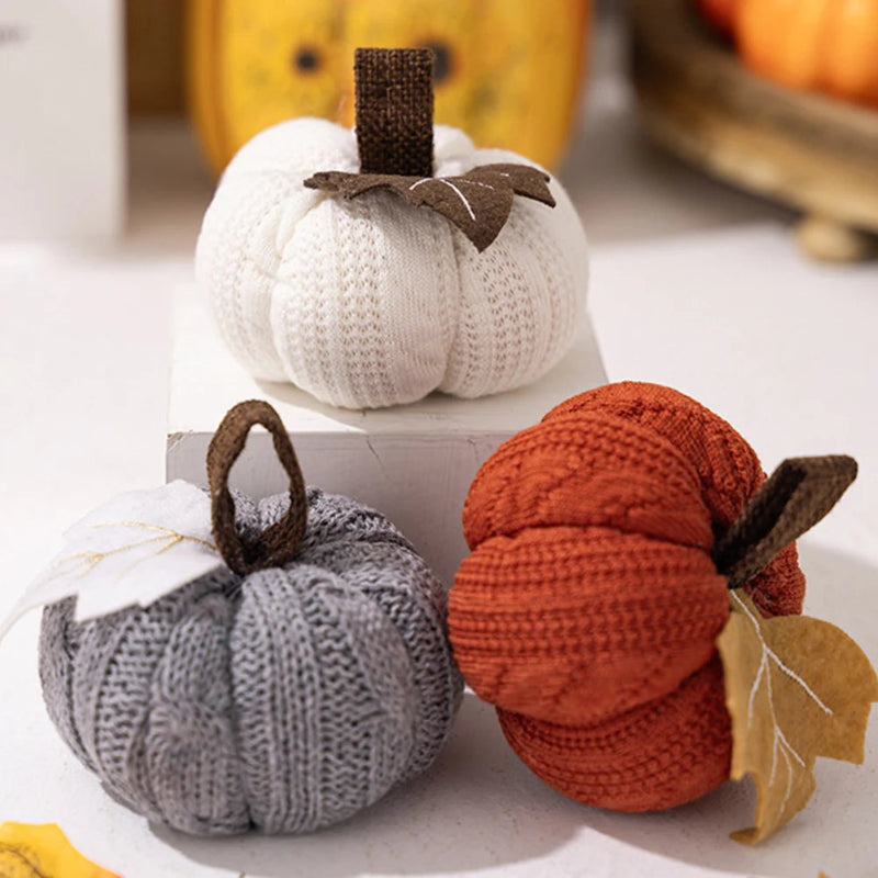 Three knitted pumpkins, in white, orange, and gray, sit on a white surface with fall leaves.