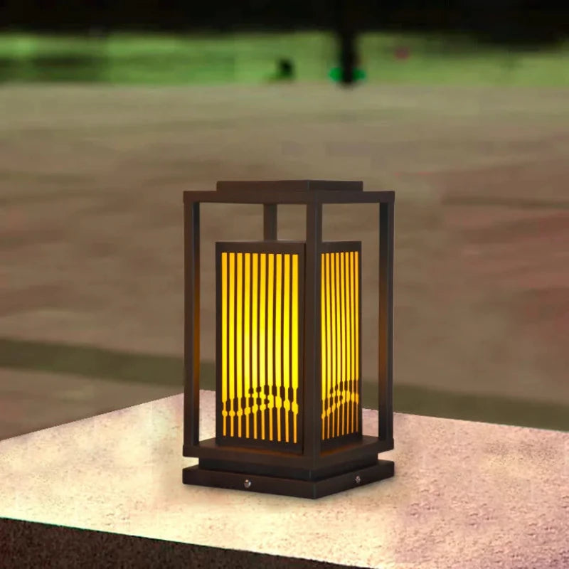 A modern, dark-framed outdoor lantern with vertical slats emits a warm yellow glow as it sits on a light-colored stone surface. A blurred outdoor background is visible.