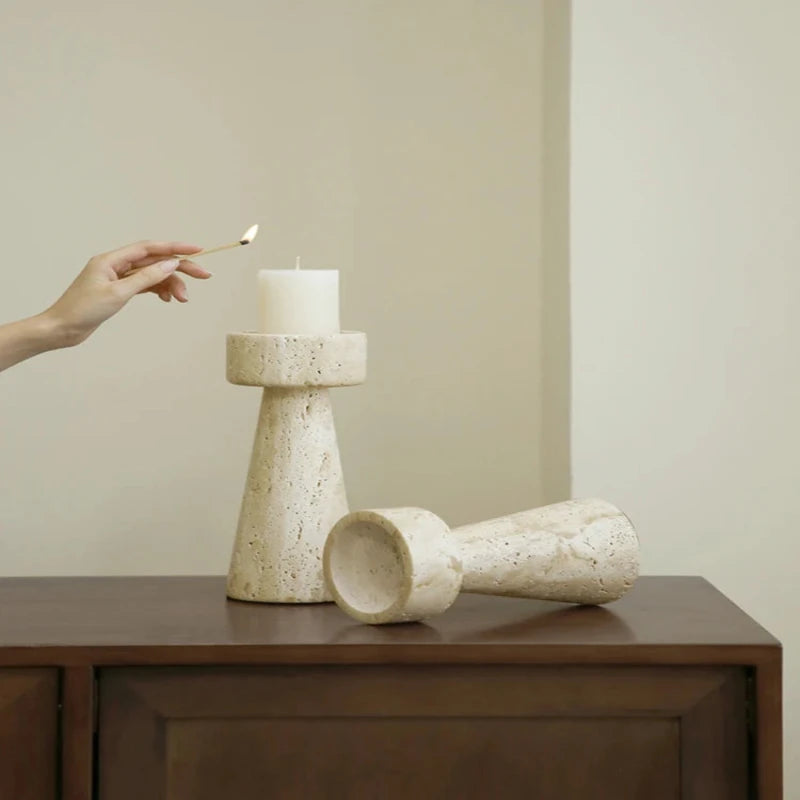 A hand holding a lit match to a white pillar candle in a textured, light-colored faux travertine stone candle holder, with another similar candle holder lying on its side on a dark wooden surface against a neutral wall.