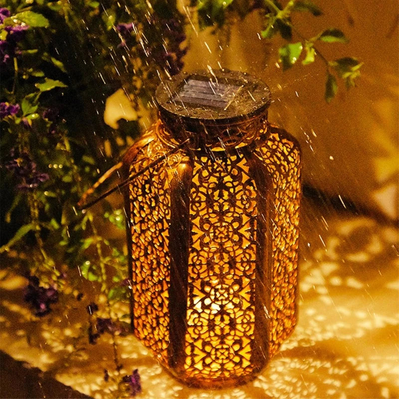 A decorative metal solar lantern with intricate cutouts emits a warm glow while raindrops fall on it. The lantern sits on a surface that reflects its patterned light and shadows. Foliage with purple flowers is visible in the blurred background.