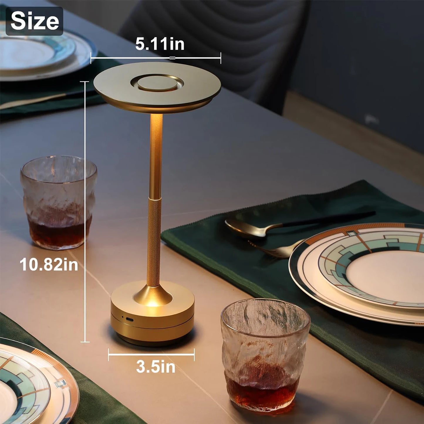 A gold-colored modern LED table lamp with a circular flat top and a textured stem, placed on a dining table. The lamp's dimensions are displayed: 10.82 inches in height, 5.11 inches in top diameter, and 3.5 inches in base diameter. The table is set with plates, green napkins, gold utensils, and two glasses partially filled with a dark liquid.