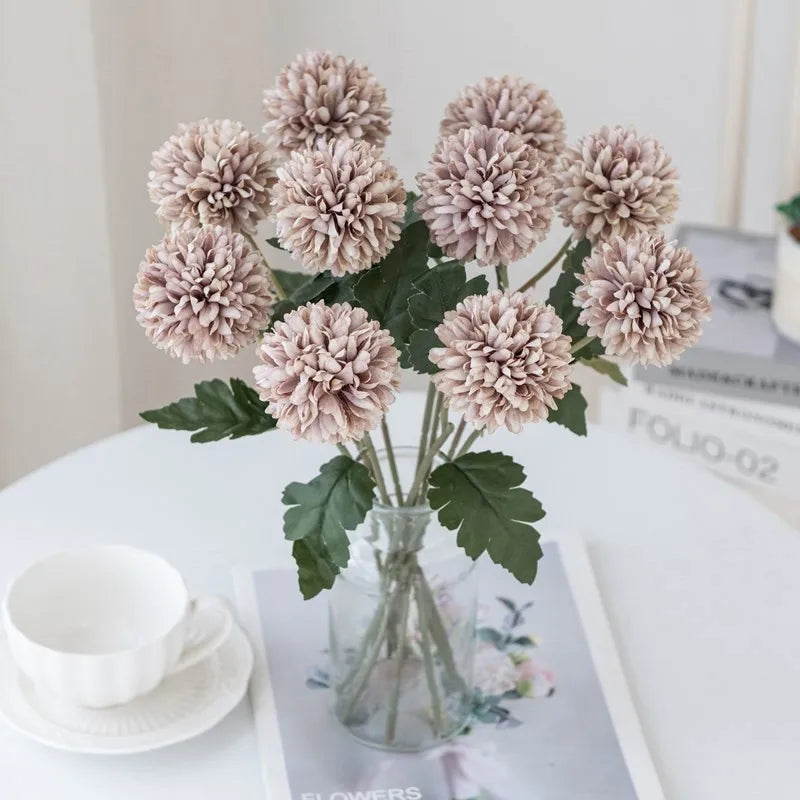 Light purple faux chrysanthemum flowers in a clear vase on a white table.