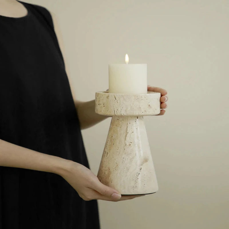 A person's hands gently holding a textured, light-colored faux travertine stone candle holder with a lit white pillar candle. The person is wearing a dark sleeveless garment, and the background is a plain light beige.