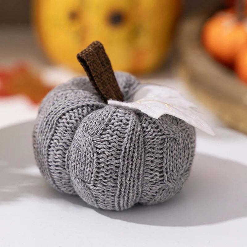 A close-up of a gray knitted pumpkin with a brown stem and a white leaf, sitting on a white surface.