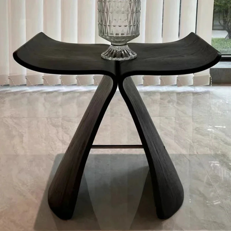 A black Butterfly Stool, serving as a small table, holds a clear textured glass vase. The stool sits on a light-colored tiled floor in front of sheer white curtains and a window.