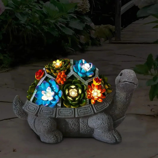 A decorative gray stone turtle statue with a segmented shell containing illuminated colorful artificial succulents is displayed outdoors at dusk or night. The lights within the succulents emit a soft glow. Dark foliage is visible in the background.