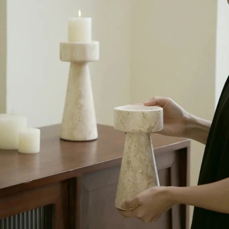 A person's hands placing a textured, light-colored faux travertine stone candle holder on a dark wooden surface next to another identical candle holder with a lit white pillar candle and two unlit pillar candles. The background is a plain light wall.