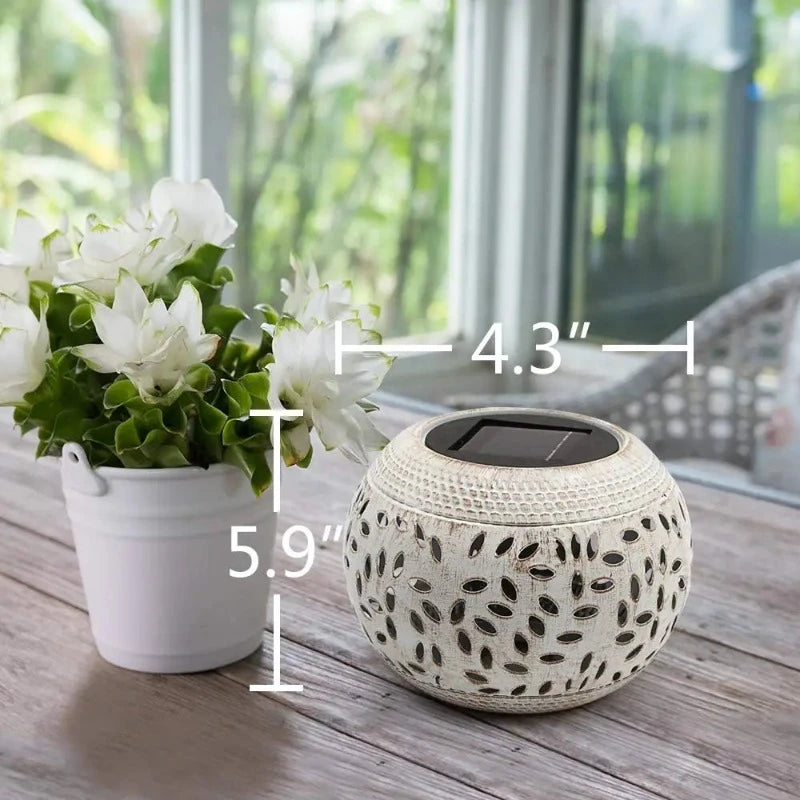A diagram shows the approximate dimensions of a round, off-white outdoor lantern with decorative oval cutouts. The lantern has a dark, flat top, likely a solar panel. An arrow indicates the approximate width of the lantern as 4.3 inches, and another arrow shows the approximate height as 5.9 inches. A small white pot with white flowers and green leaves sits to the left on a wooden surface. A blurred background shows a window and greenery.