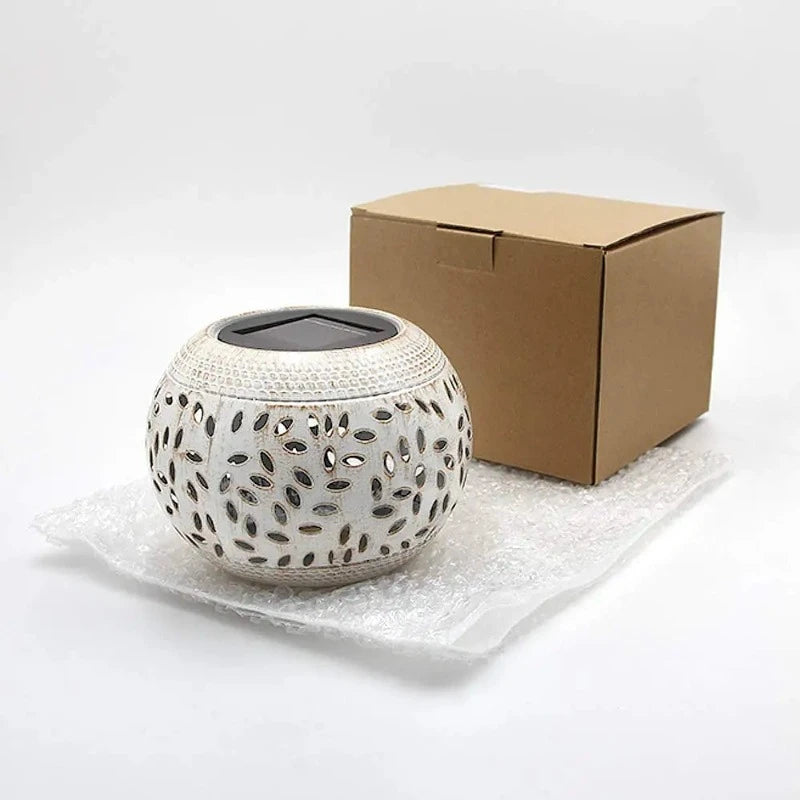 Okay, here's an accurate, detailed, and shorter alt text option for your Shopify, avoiding brand mentions:
A round, off-white outdoor lantern with decorative oval cutouts sits on a light-colored surface with a cardboard box in the background. The lantern has a dark, flat top, likely a solar panel.
