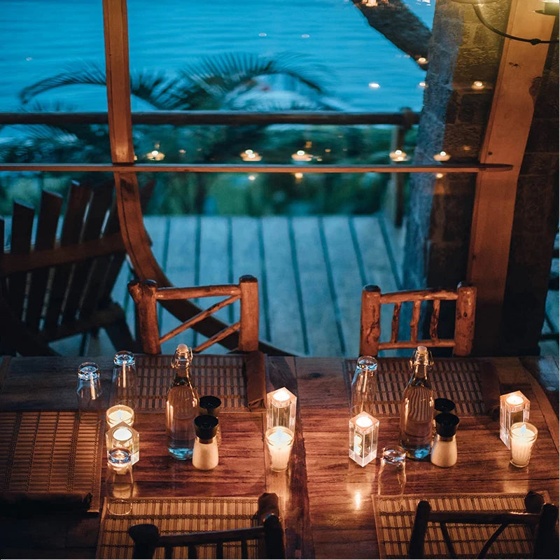 A rustic wooden dining table is set for a romantic outdoor dinner overlooking a dark ocean at twilight. The table is illuminated by several small, glowing candle holders. Clear drinking glasses, bottles, and salt and pepper shakers are also arranged on the table. Wooden chairs are placed around the table, and string lights twinkle in the background, enhancing the intimate ambiance.