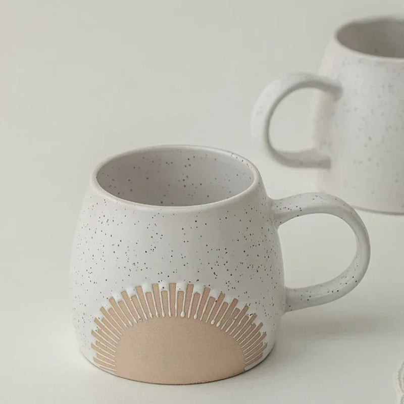 A speckled ceramic mug with an embossed sunburst design sits on a light-colored surface, with another similar mug slightly out of focus in the background. The mug has a smooth curved handle and a modern, minimalist aesthetic.