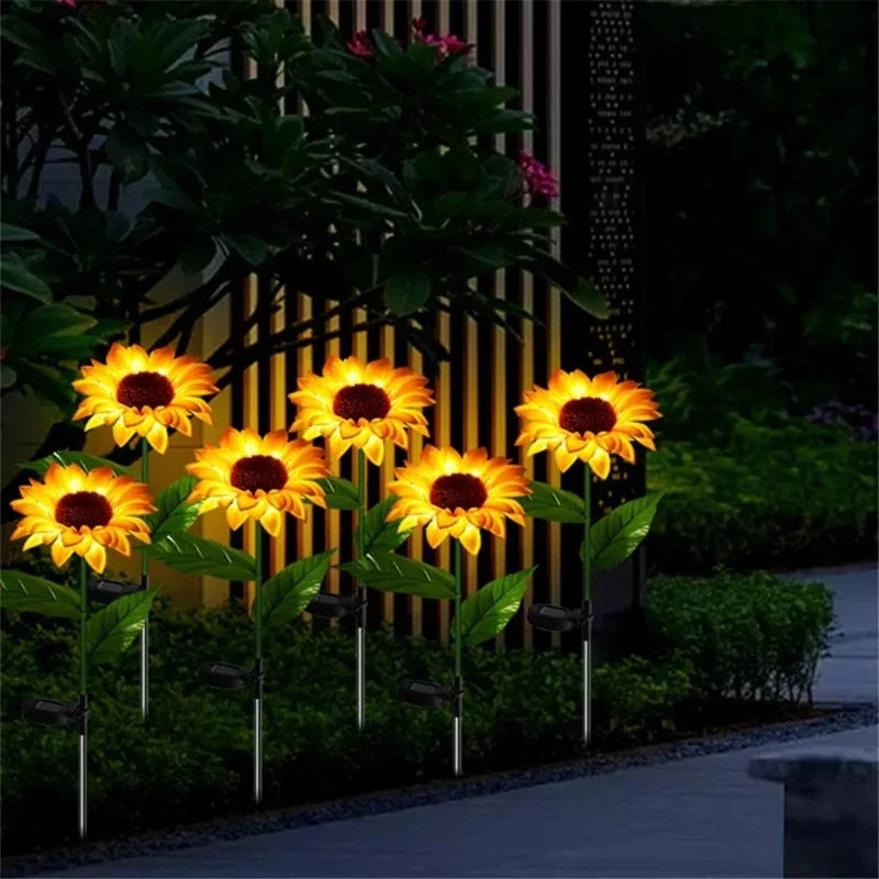 Six artificial sunflowers with dark centers and warm yellow glowing petals are planted along a garden path at night. Each flower is illuminated and has green leaves and a small black solar panel near its base. The background shows dark foliage and a fence with vertical slats. The scene depicts an illuminated garden walkway.