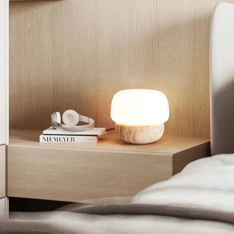 A small, glowing table lamp with a rounded white shade and a textured, light-colored base sits on a light wooden nightstand. A pair of white headphones rests on top of a book next to the lamp. The scene is next to a bed with white linens and a textured grey blanket, against a light wooden headboard.