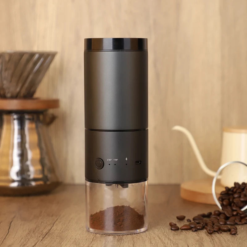A sleek black electric coffee grinder with a compact, modern design, featuring a transparent lower chamber filled with freshly ground coffee. The warm wooden background and surrounding coffee accessories create a cozy and inviting coffee-making ambiance.