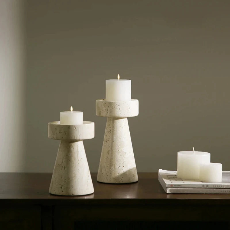 Two textured, light-colored faux travertine stone candle holders of varying heights, each holding a lit white pillar candle, arranged on a dark surface with two unlit white pillar candles resting on a stack of magazines.