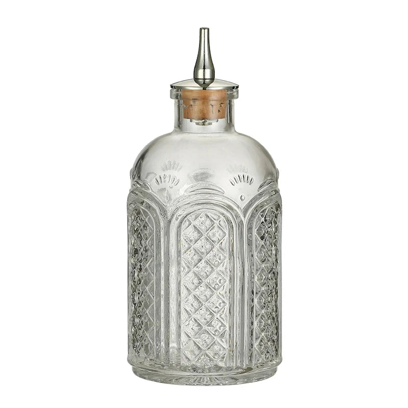 This bottle features a unique vintage design with intricate embossed patterns, including diamond lattice textures framed by arched borders. The upper portion has subtle decorative flourishes. The bottle has a rounded shoulder leading to a narrow neck, topped with a cork stopper and a metal spout for controlled pouring. The clear glass enhances its elegant aesthetic.